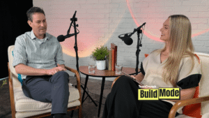 A podcast-style interview setup shows a man and a woman seated across from each other in armchairs, speaking into microphones. They sit beside a small round table with water glasses, books, and a potted plant, against a white brick wall backdrop. A “TechCrunch Build Mode” logo appears in the lower corner.